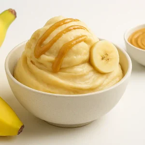 طرز تهیه بستنی موز و بادام‌زمینی در یک کاسه سرامیکی مینیمال با تزئین برش موز و ریزش کره بادام‌زمینی، همراه با موز کامل و کاسه کره بادام‌زمینی در پس‌زمینه سفید و خلوت